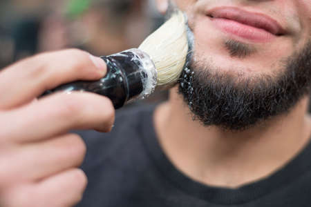 Closeup of a barber putting some shaving cream on a client's face before giving him a shave shaving a beard to a caucasian man. Lathers a beard, preparing it for a haircut. Barber Shop Professional Beard Careの写真素材