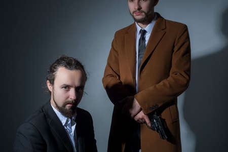 Portrait of two men in business suits, one of them is sitting, the other is holding a gun. On a gray background. Dramatic light. Artistic setting. The concept of criminal authorities. Businessmen bandits. Disassembly businessmenの写真素材