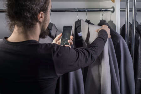 Close-up portrait of a thousand-year-old young bearded guy in a men's clothing store, chooses a business suit for work, photographs a high-quality jacket on a smartphone. Sale of men's clothing. business jacket. Gray and beige business suit. A man touches suits, checking the quality of clothing fabricの写真素材