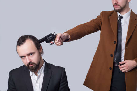 business man in a suit aiming a gun at the head of another man in a white shirt. on a gray...