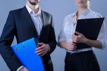 Portrait of young people, businessmen smiling, stand in business suits, holding office folders for papers in their hands. Studio photo on a gray background. Simple business concept. Happy office work. Office clerk. Serious business portraitの写真素材