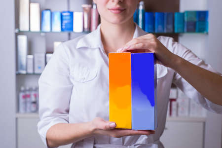 Closeup of a defocused girl, a cosmetologist, a specialist in the sale of cosmetics for body care, keeps the package in focus. showing off makeup. In the background is a counter of cosmetics for the care of face skin, hair, means of treating and restoring the life of skin tissues, facial cleansing, a stimulant for the faceの写真素材