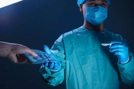 Portrait of a surgeon, in a sterile gown, hat and mask. Holding a scalpel in one hand, he takes a press of dollar paper money in the other hand. Surgical concept .Advertising medical surgical photo portrait for a magazine. On a white background Against the background of an operating lamp. Studio photo medicine. Operating room. Modern methods of treatment in surgery. Procurement for innovative surgery topics in the world. Free space. Bribe in medicine, the fight against bribery in medicineの写真素材