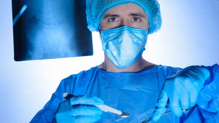 Portrait of a surgeon in sterile gowns, hats and masks. Holding a scalpel and a needle holder with a...
