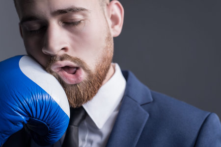 close-up portrait of a young bearded guy in a business suit, businessman, side view the hand of an opponent in a boxing glove strikes.の写真素材