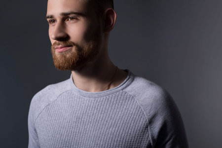 closeup, dramatic portrait of a handsome bearded guy of twenty-five years old, in a gray sweater, on a gray background. Casual style. Sweater, mens jacket. Smiling bearded young guy. Dark colored man portraitの写真素材
