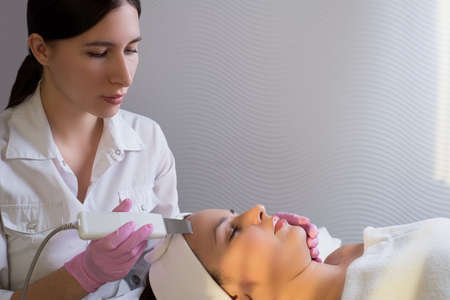 close-up, professional beautician using an ultrasonic facial cleanser, cleans the skin of a girl patient, in a beauty salonの写真素材