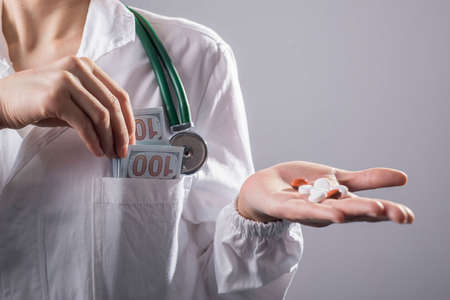 Stethoscope Laying on Stacks of Hundred Dollar Bills with Narrow Depth of Field.Close-up of a doctor in a white coat, with a phonendoscope, in his pocket a press of dollar bills, and a doctor's hand pulling money out of his pocket. On a gray background, studio photo. Bribe conceptの写真素材