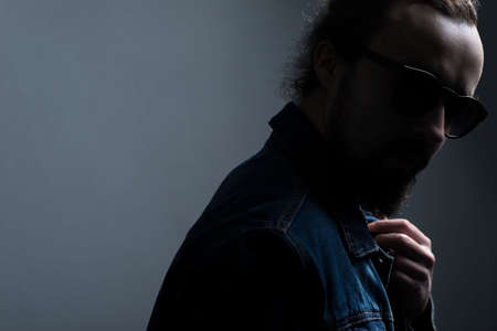 Studio dramatic shadow portrait of a young bearded guy with glasses, in a denim jacket, on a gray...