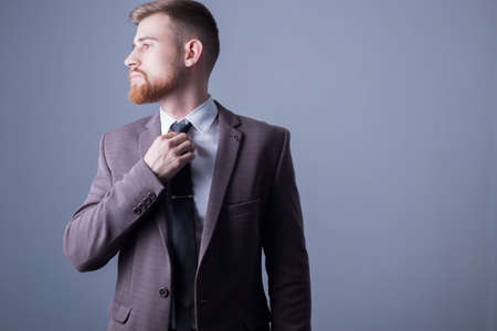 Studio portrait of a young bearded handsome guy of twenty-five years old, in an official suit, tightens his tie. On a dark backgroundの写真素材