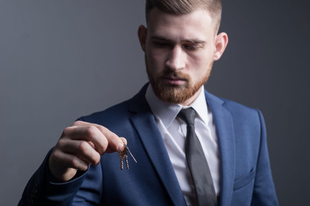 close-up portrait of a young bearded guy of twenty-five years old, in a business suit, holding keys in his hand. On a gray background. conceptの写真素材