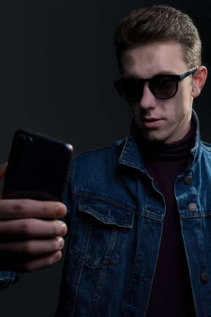Closeup portrait of a young guy, in sunglasses, holding a smartphone, taking a selfie. Denim style. on a gray backgroundの写真素材
