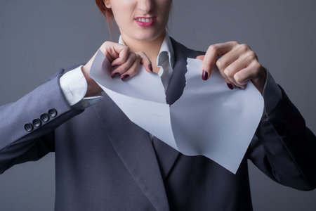close-up. Studio portrait of a young business woman, with red lipstick, and a gray business jacket, tearing a blank paper, with a grin. On a gray backgroundの写真素材