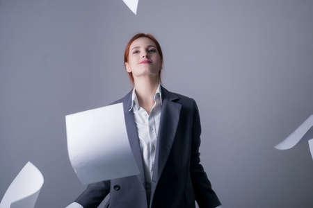 young red-haired business woman, with red lipstick, and a gray business jacket, holding many blank sheets of paper in her hands, scattering themの写真素材