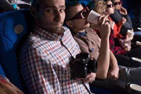 a young guy, looking around, in fear of being seen behind an illegal pirate video in a movie theater. Against the backdrop of sitting people of different sexesの写真素材
