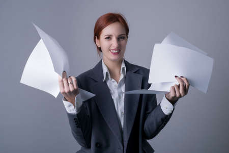 portrait of a young red-haired business woman, with red lipstick and a gray business suit, holding in her hands a lot of blank sheets of paperの写真素材