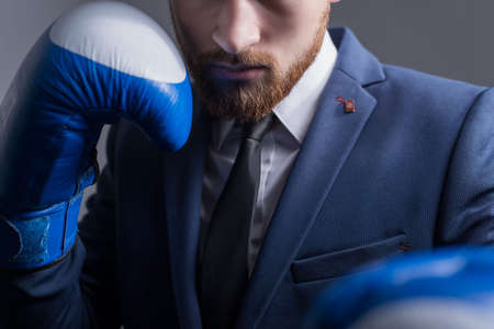 main plan, studio dramatic portrait of a handsome young bearded guy of twenty-five years old, a man in a business suit, looking at the camera, holding boxing gloves on his face. On a gray background. Business concept. Hold the punch. The guy looks like Conor McGregor. Business bearded guy. Sport and business. A man in a tuxedo. The official style of men.の写真素材