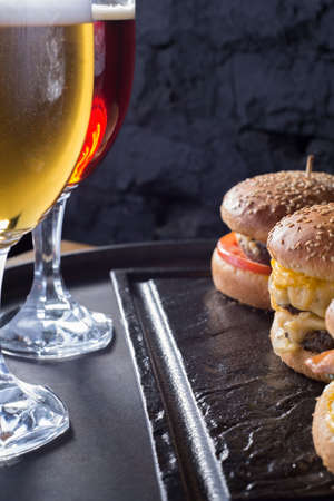 mug of beer with hamburger and fries close-up, beer glasses with craft dark and light beer with foam, against a background of burgers, with a variety of ingredients, including shyomga, beef meat patty, cheese burger. In a rustic style, against a black brick wall background. Junk food. Alcoholic drink. Tasty foamy draft beer of a dark grade. delicious burger. Promotional photo for a beer burger menuの写真素材