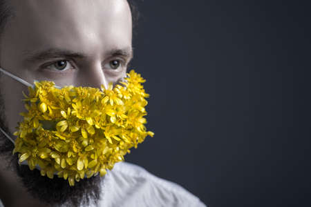Close-up, studio portrait of a young guy, in a medical mask of flowers, looking to the side. On a gray background. Medical concept of coronavirus infection. Protection against colds by masking the airways. Spring concept of allergic diseases, with a background on allergies from pollen of flowers. Mask on the face of flowers. Allergic Disease Treatmentの写真素材