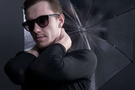 Dramatic studio portrait of a guy in black, holding an umbrella in sunglasses. On a gray background. Hide from the rain under an umbrella.の写真素材