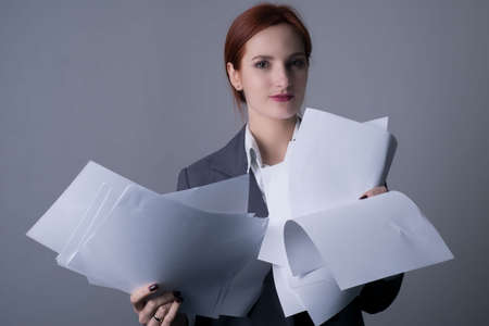 close-up. Studio portrait of a young red-haired business woman, with red lipstick, and a gray business jacket, holding many blank sheets of paper in her hands. On a gray background.の写真素材