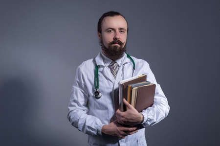 Portrait of a bearded doctor in a white coat and a phonendoscope holding educational books in his hands. Studio photo on a gray background.の写真素材