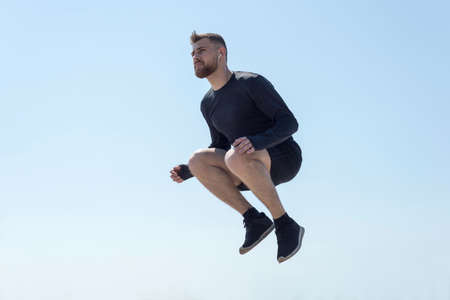 Portrait of a young bearded athlete of twenty-five, jumping against a blue sky athlete running along...