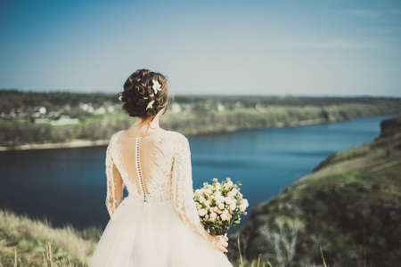 general plan portrait from the back of a young bride, standing with a wedding bouquet, looking into the distance of a landscape with a river. Creative art tinting. Wedding photo of a young beautiful bride of twenty-five years, waiting for the holiday. Single brideの写真素材