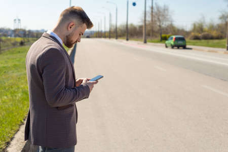 Using cellphone outdoors - with defocused city traffic. Portrait of a young bearded guy of twenty-five years old, using a smartphone, stands at the carriageway, against the background of a car. A man calls the rescue service, emergency service using the smartphone applicationの写真素材