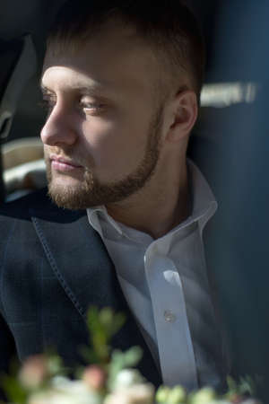close-up portrait of a young bearded groom, twenty-five years old, with flowers for the bride, riding in a car, looking out the window. Waiting for the groom before the wedding. Wedding taxi, the groom in the car rides for the bride. Dramatic wedding portrait of the groomの写真素材
