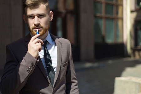electronic cigarettes . Portrait of a young bearded guy of twenty-five years old, stands by the textured wall of the building, smokes a cigarette.の写真素材