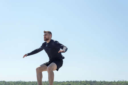 Portrait of a young bearded athlete of twenty-five, jumping against a blue sky athlete running along...