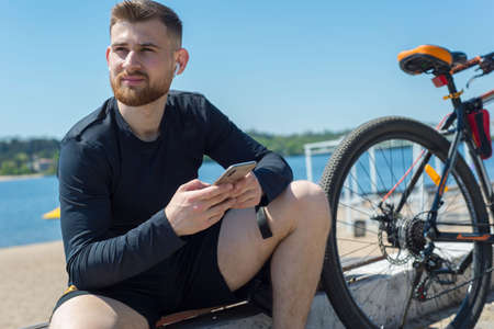Portrait of a bearded cyclist athlete guy resting after a bike ride using a smartphone in his hand. Outdoors. Spring sports. The guy is twenty-five years old. Cycling Spring bike rideの写真素材
