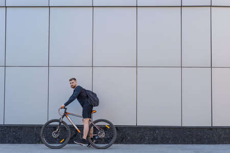 bearded cyclist on a bicycle on a gray building background. Outdoors. Spring sports. The guy is twenty-five years old. Cycling Spring bike ride. Extreme cycling. Active lifestyle.の写真素材