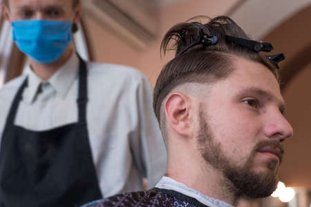 a barber in a protective mask in a barbershop is cutting a young bearded guy. quarantine haircut, barbershop coronavirusの写真素材