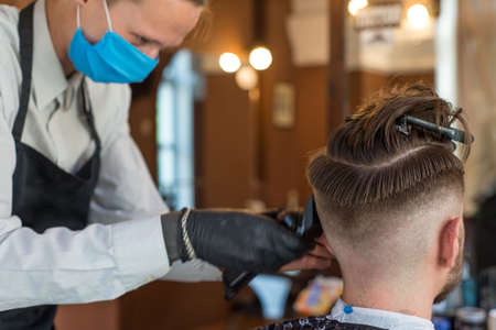 a hairdresser cuts a bearded young guy with a hair trimmer, combing the hair on his head. Work of...