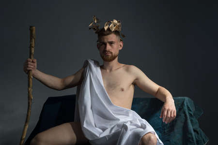Portrait of the ancient Greek god Olympus, in white clothes, with a wreath on his head, holds a staff, sitting on a dark bench.の写真素材