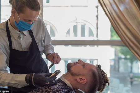 a barber in a protective mask in a barbershop is cutting a young bearded guy. quarantine haircut, barbershop worker in a protective mask shaving a guy, men shaving facial hair, barbershop in the epidemic of coronavirus infection, hairdresser service worker The work of the barber during the coronavirus, the hairdresser trim the client in a mask and a protective suit , quarantineの写真素材