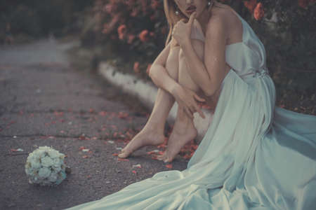 portrait of a lonely bride, in a white dress, sitting on the pavement, against the background of a floral background of rosesの写真素材