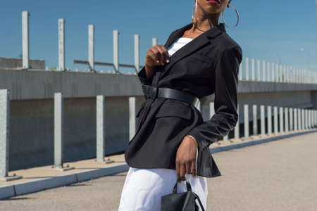 Fashionable African girl in a business jacket, and a white dress, holds a backpack in his hand. Fashionable advertising shoot against a blue sky. A young black African girl, in a black business formal jacket, over a white dress. A new fashion for wearing a belt at the waist over a female business black jacket. Relkma of a female leather backpack close-up. Beautiful black girl with a white dress posingの写真素材