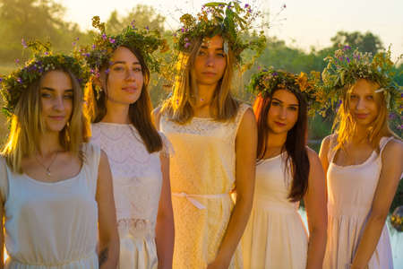 a group of young girls of twenty years old, in white dresses, with wreaths of flowers, hugging, standing against the sunset at the pond.の写真素材