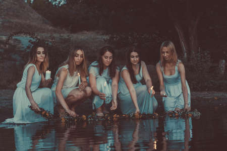 a group of young tangled girls in white dresses, launching flower wreaths in the water hold candles. On a natural background. A rustic style, a national holiday for young bridesの写真素材