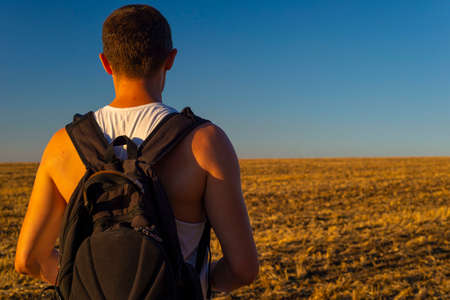 tourist young guy with a backpack, standing with his back, looks into the distance of a wheat field....