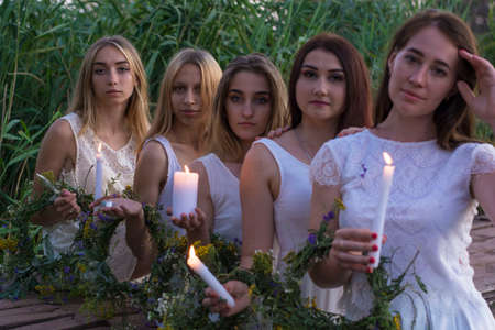 a group of young tangled girls in white dresses, launching flower wreaths in the water hold candles. On a natural background. A rustic style, a national holiday for young brides of Russia and Ukraineの写真素材