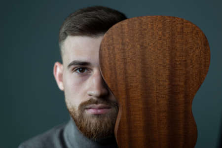 a portrait of a twenty six year old bearded young man covering half of his face with a ukulele. On a...