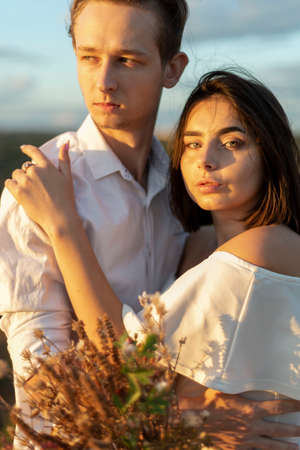portrait of a young beautiful couple, girl with a guy with flowers. Standing in white clothes in the field. Against the backdrop of the setting sun, summer evening, cloudy blue skyの写真素材