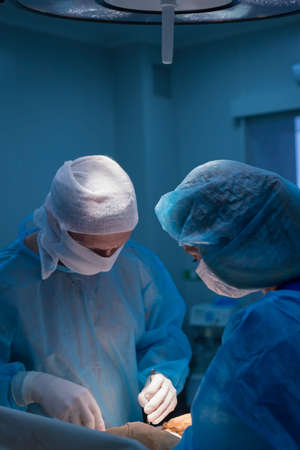 Children's surgeons perform urological operations. A man and a woman in a mask and a blue sterile coat in the operating room. A team of surgeons is conducting an operation. Treatment of inguinal hernia, testicular edema, testicular disease, varicocele, cryptorchidism. Focus on the warm light of the lamp, the background is illuminated by cold blue light.の写真素材