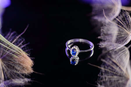 white gold ring with sapphire stone, against a black mirror background, in soft pastel violet tones. With a dandelion flower. Jewelryの写真素材