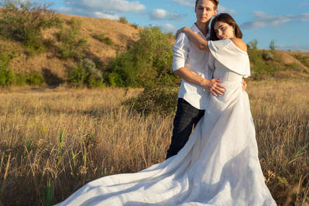 young wedding couple of a guy and a girl, in a white dress, standing in a field, against the backdrop of a cloudy blue sky, sunset. Love of young people. generation z. Natural photo in the field of a wedding couple, young people twenty two years old. A newly wed couple walking through a grassland, holding handsの写真素材
