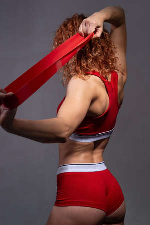 studio portrait of a young athlete of an athletic physique, in red, stretching a fitness elastic band. On a gray background Hands of a young sporty woman with dreadlocks pulling red elastic band in the park. In sportswear. Sports concept.の写真素材